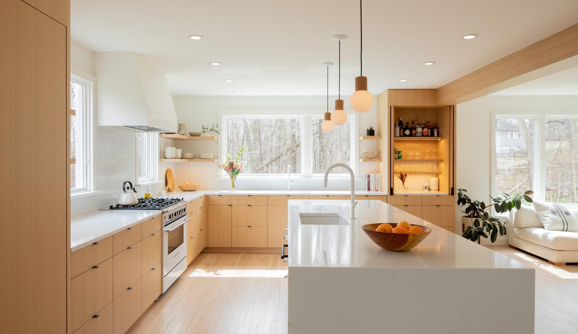 A spotless kitchen with light oak cabinets, a white quartz island, pendant lights, and a bowl of oranges