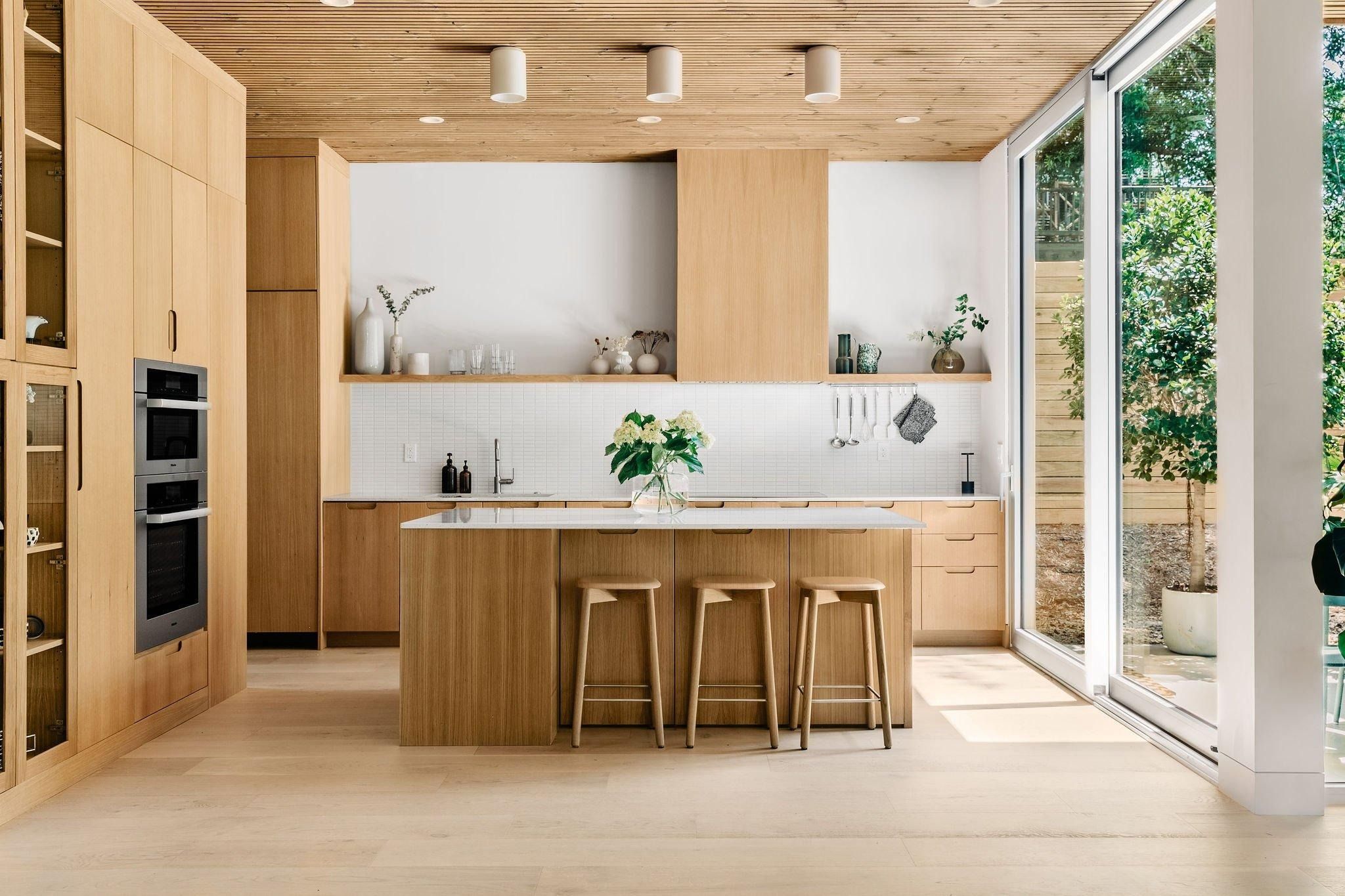 A kitchen with a slatted wood ceiling, oak cabinets, and a glass wall open to a sunny garden